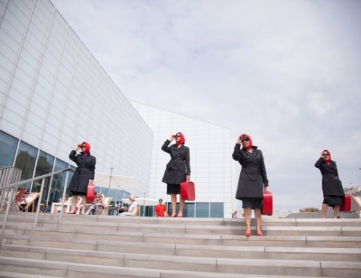 A photo of some ladies in black coats, sunglasses, red headscarves and red briefcases standing on the steps outside The Turner Contemporary Gallery